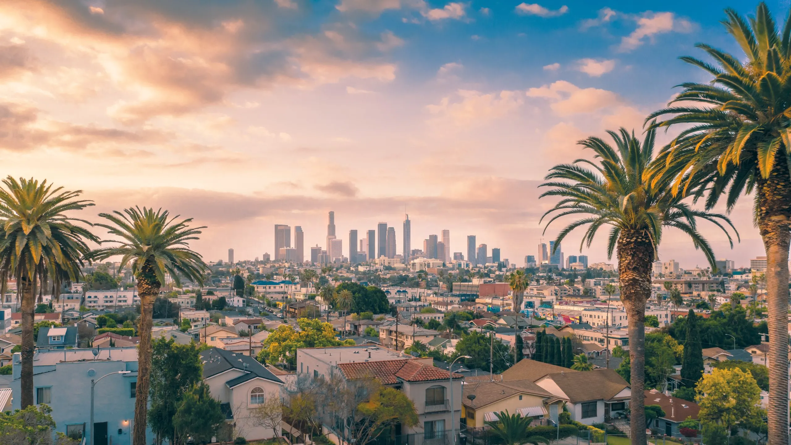 Sunset view of Los Angeles with downtown skyline, palm trees, and Mediterranean-style rooftops under a pink and orange sky.