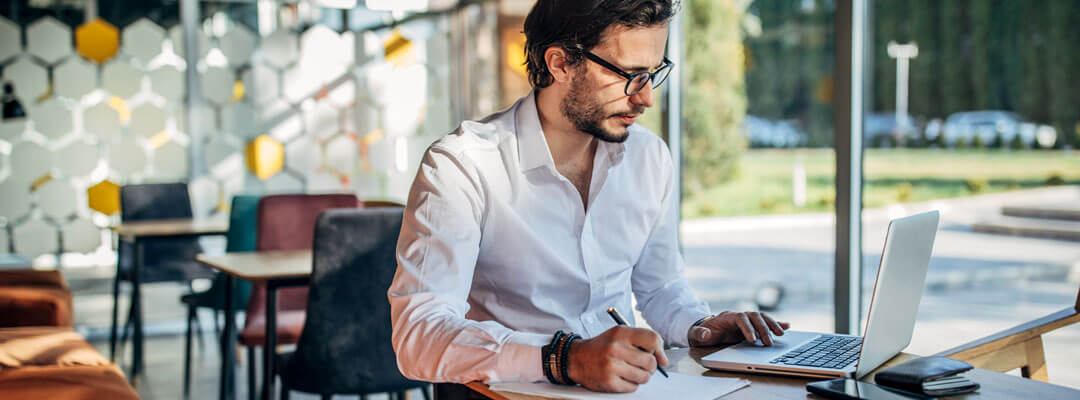 A man scrolling on his laptop while writing down on a paper on a cafe table on Southern California Cafe