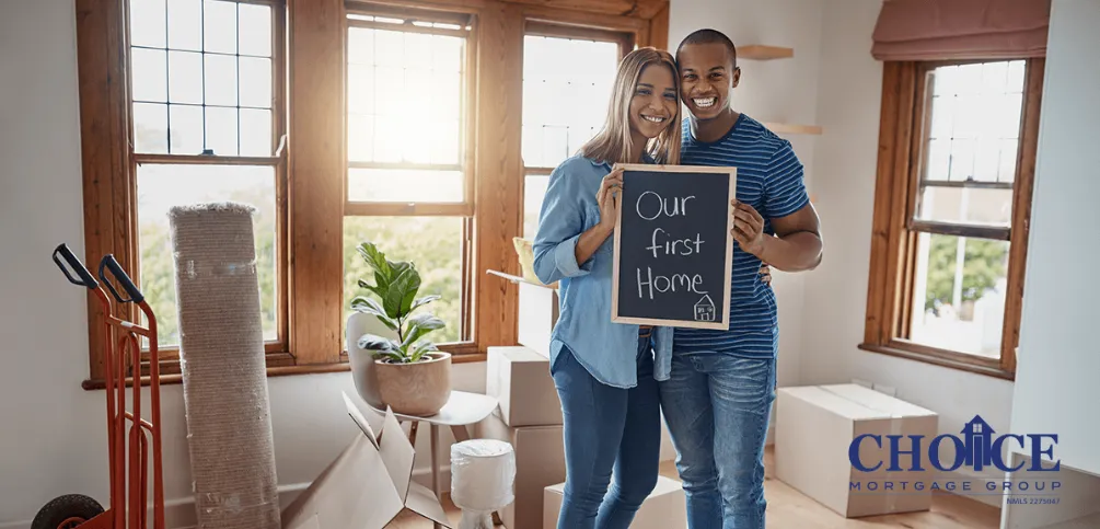 Two young couple holding a signage written "Our first home" with boxes of around them.